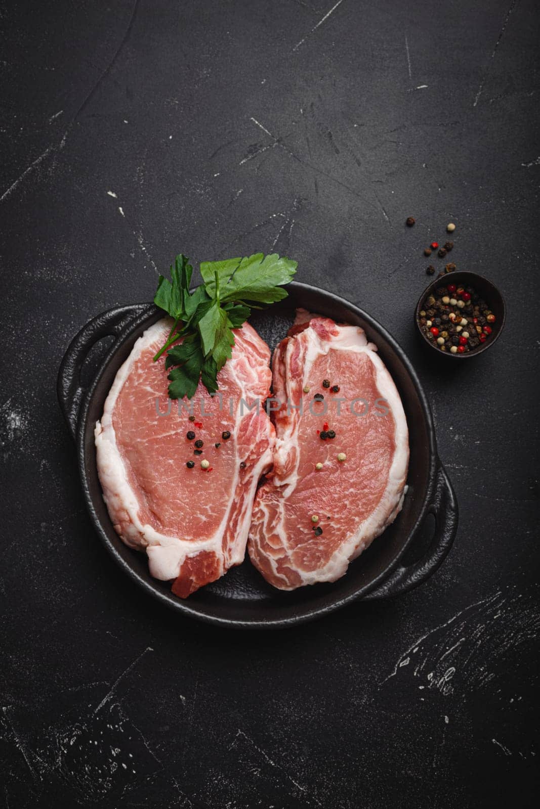 Cut raw meat pork steaks with seasonings in black cast iron pan on dark rustic stone background top view, ready for roasting. Pork loin chops cooking by its_al_dente