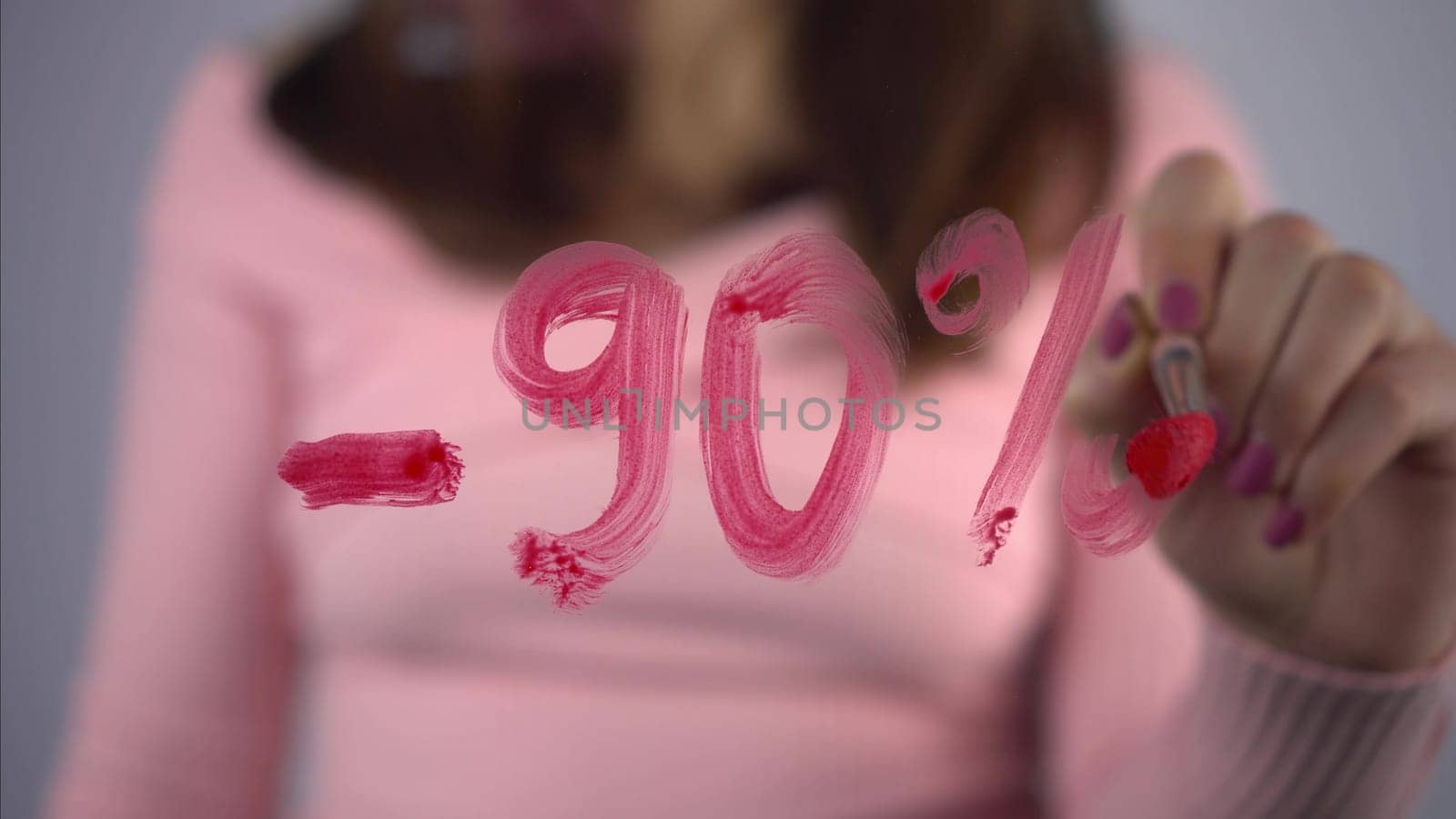 A young woman draws on the glass discounts minus 90 percent. A girl in a pink sweater draws with a brush and red paint. Close-up. 4k