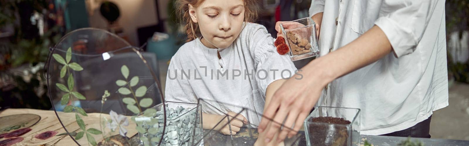 caucasian confident happy florist is working with her young daughter and making composition from glass stones and plants in botanic shop by Yaroslav_astakhov