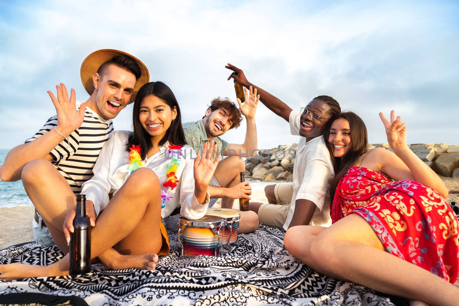 Happy, smiling group of multiracial having fun at the beach drinking beer together waving hand at camera. Vacation and friendship concept.