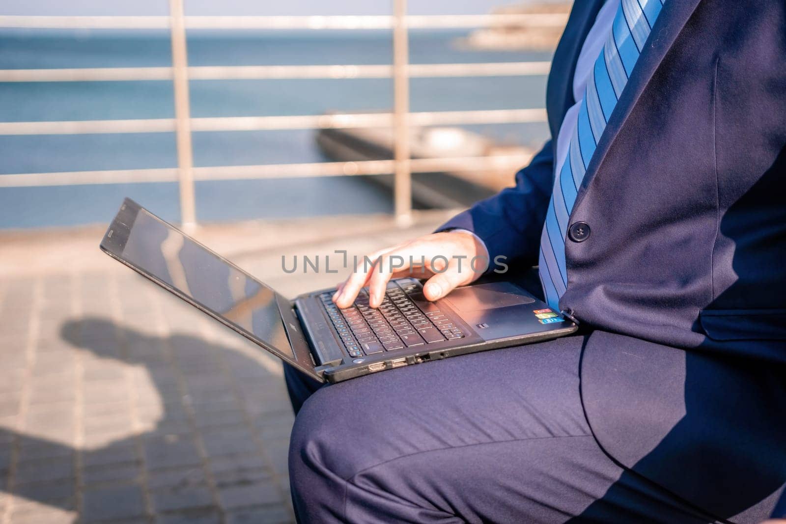 Confident middle age businessman working remotely online, typing on a laptop keyboard while sitting on a beach at sunset. Working remotely on vacation, running an online business from a distance. by panophotograph