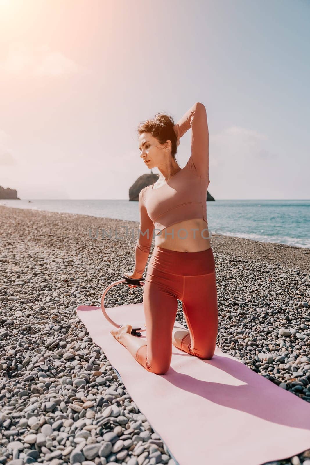 Middle aged well looking woman with black hair doing Pilates with the ring on the yoga mat near the sea on the pebble beach. Female fitness yoga concept. Healthy lifestyle, harmony and meditation.