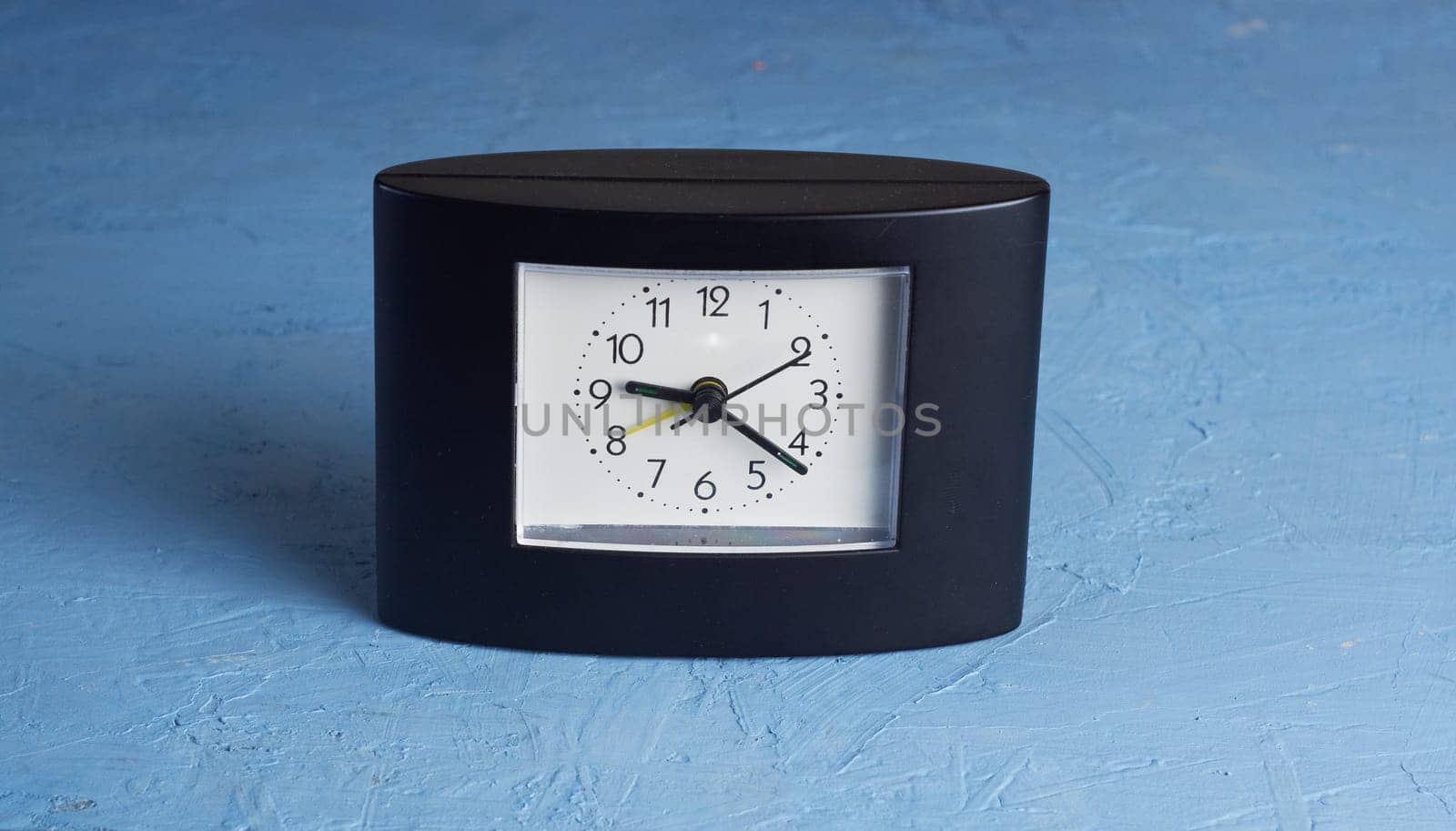 Black square-shaped desk clock with a white dial on a light blue background