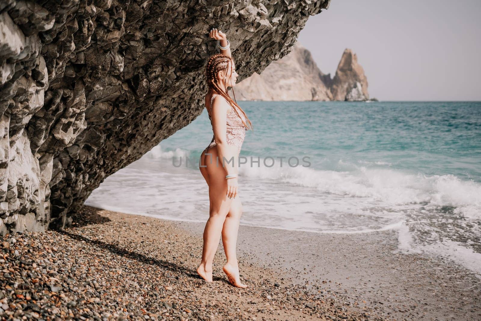 A tourist woman in bikini on beach enjoys the turquoise sea during her summer holiday. Beach vacation. Woman with braids dreadlocks standing with her arms raised near basalt rock enjoying beach ocean by panophotograph