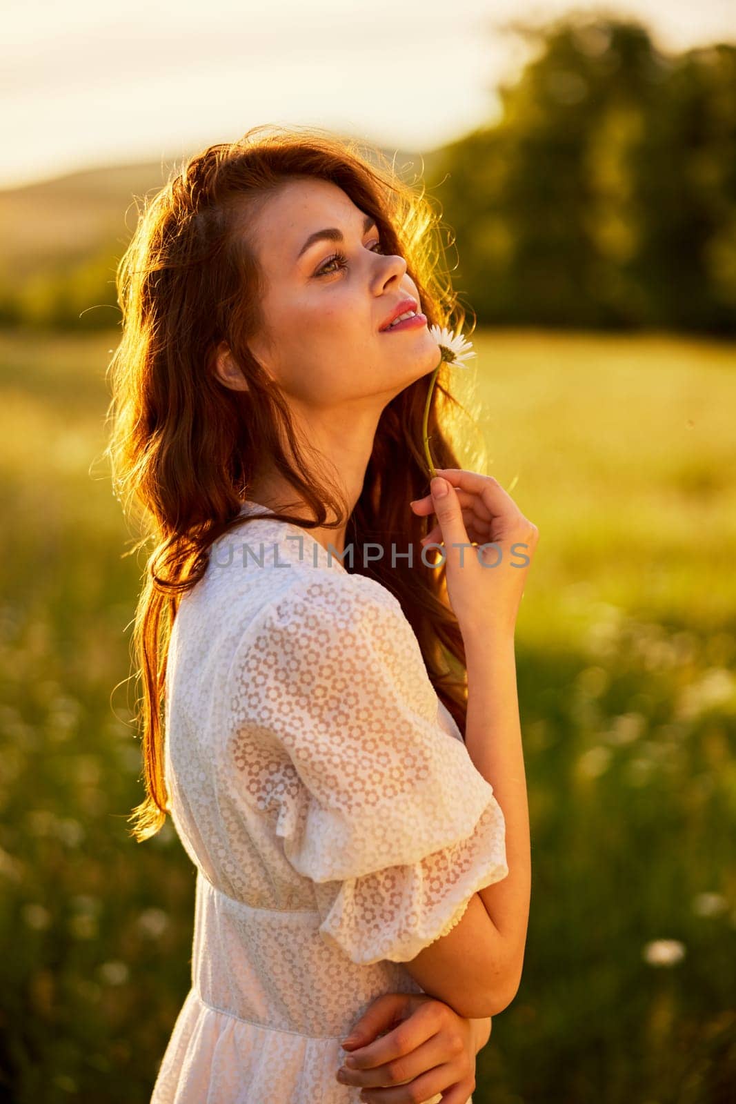 close portrait of a beautiful, laughing woman in a light dress holding a chamomile near her face during sunset, lit from the back by Vichizh