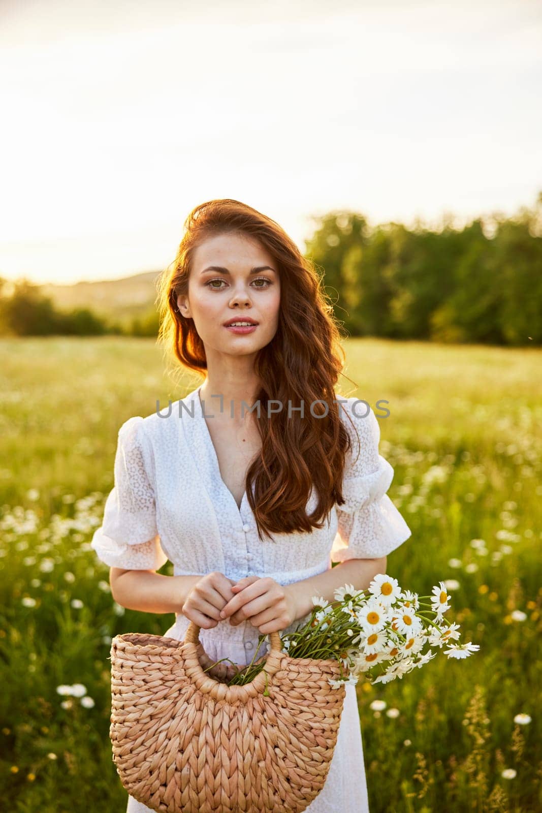 close portrait of a happy woman looking at the camera in a light dress and a wicker basket in her hands with chamomile flowers in nature. High quality photo