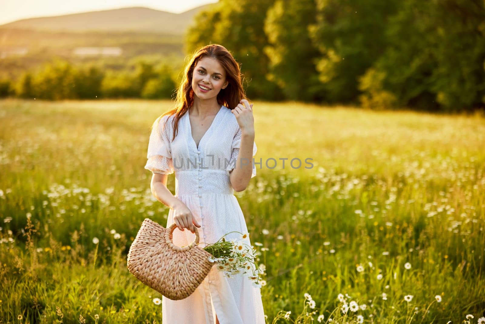 a beautiful woman in a light dress and a basket of daisies in her hands stands in a field during sunset and smiles at the camera. High quality photo