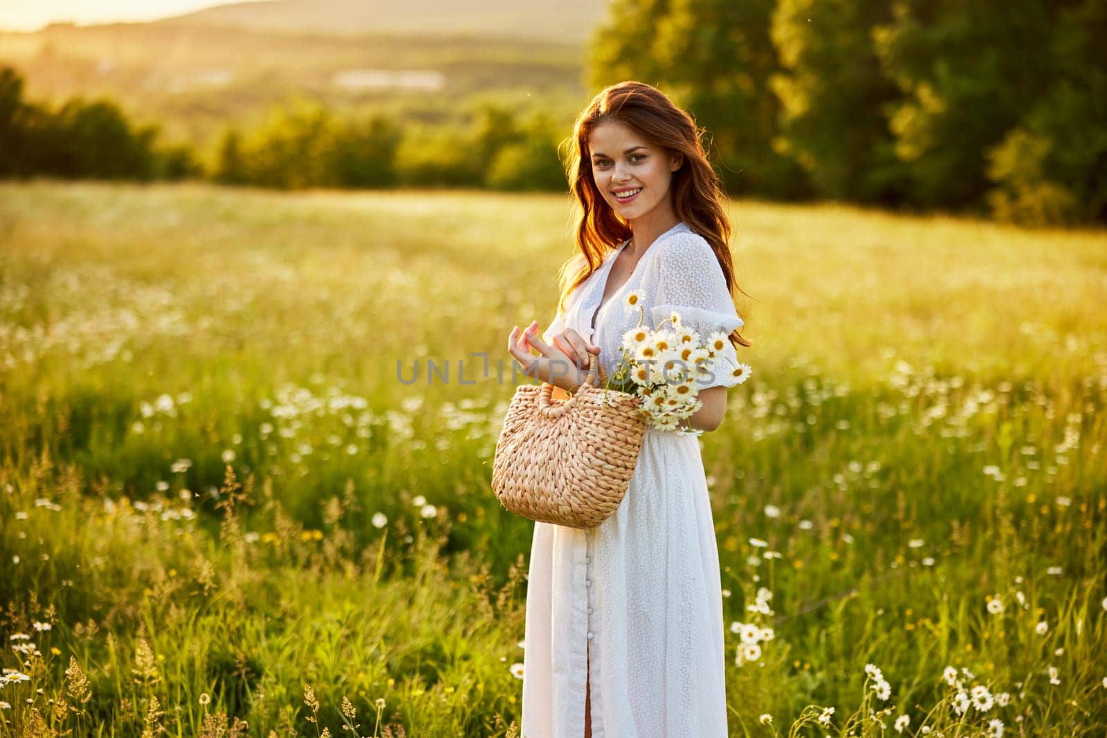 a beautiful woman in a light dress and a basket of daisies in her hands stands in a field during sunset and smiles at the camera. High quality photo