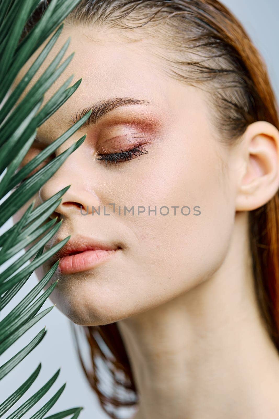 a close beauty portrait of a beautiful woman standing holding a palm leaf near her face, closing her eyes. Vertical photo without retouching of problem skin. High quality photo