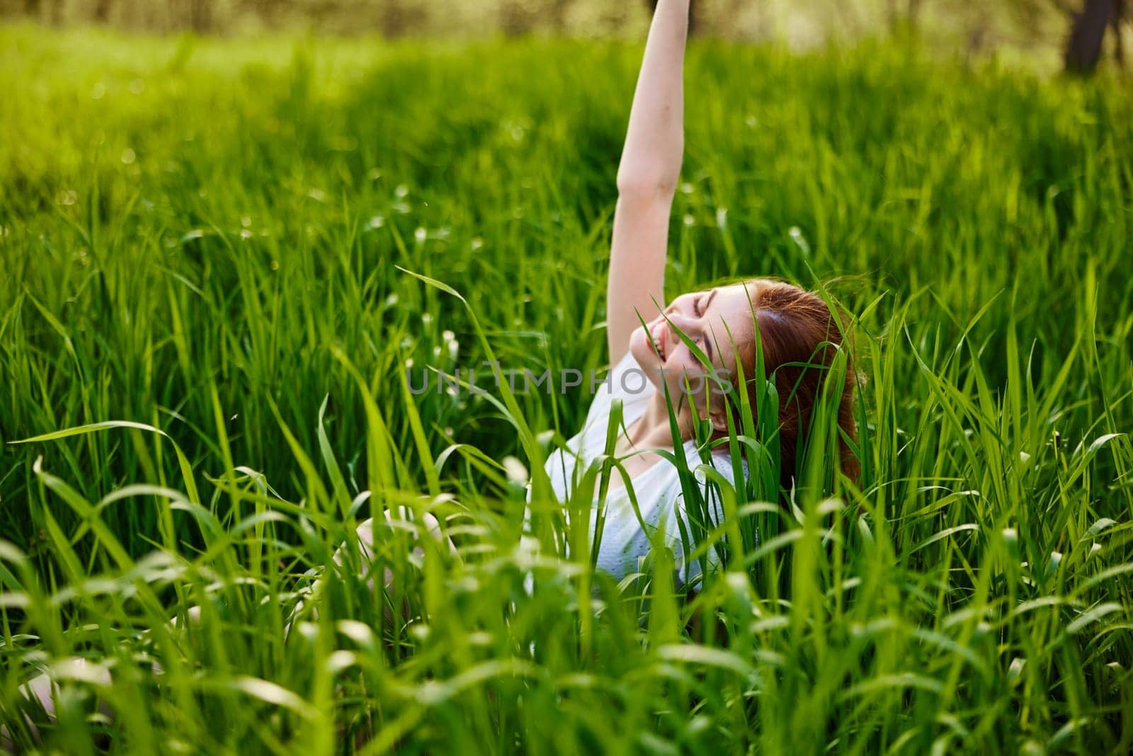 a cute woman in the summer high grass sits in a light dress happily raising her hand up. High quality photo