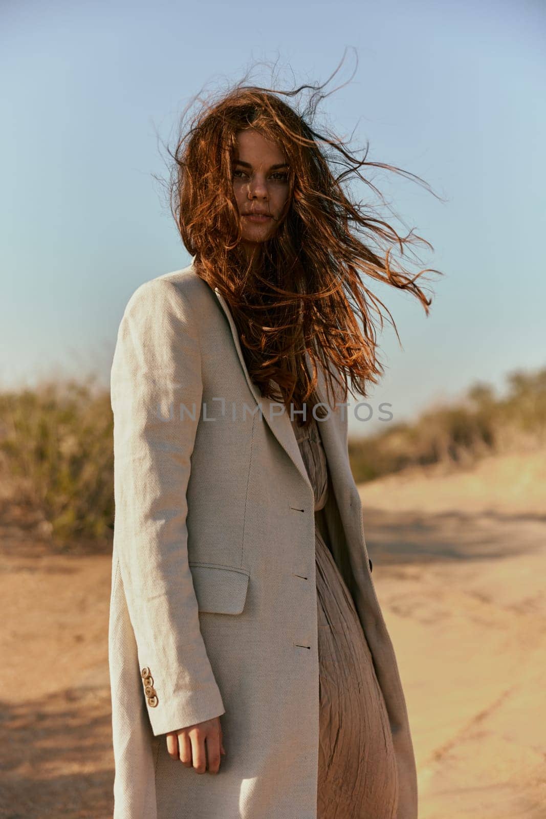 a beautiful woman in a light jacket looks at the camera while standing in nature and the wind blows her hair. High quality photo