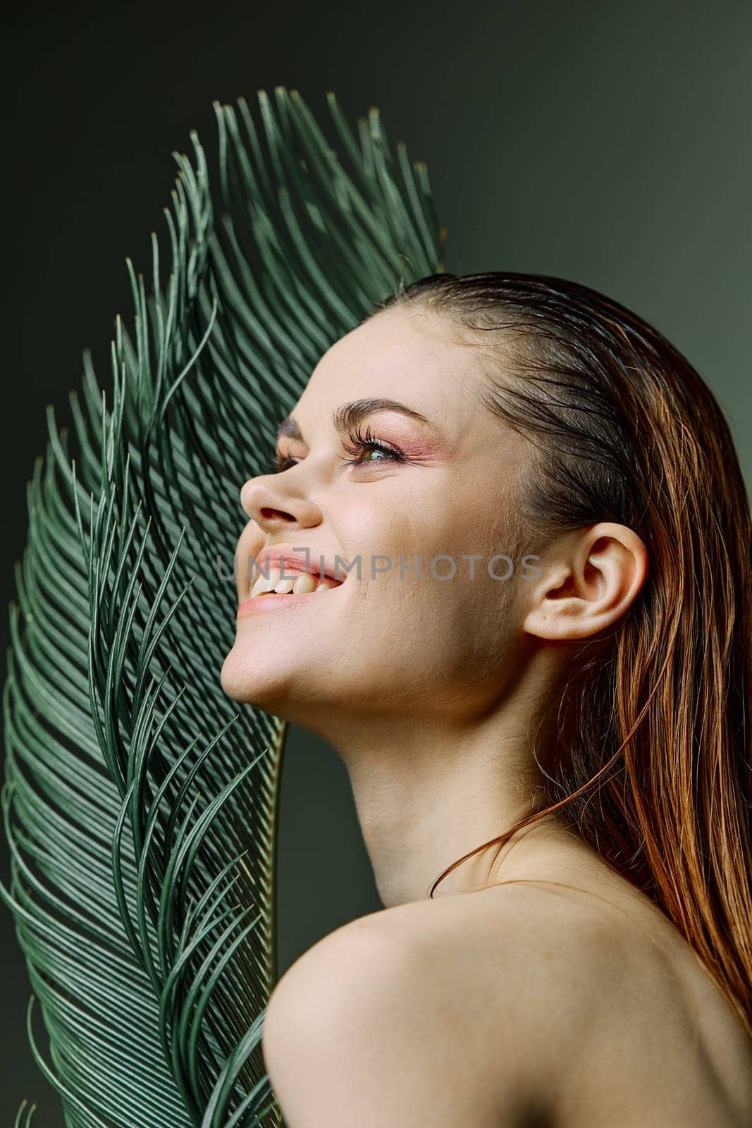 a refined, elegant woman stands with a green palm leaf holding it near her face, smiling pleasantly. Vertical photo without retouching. High quality photo