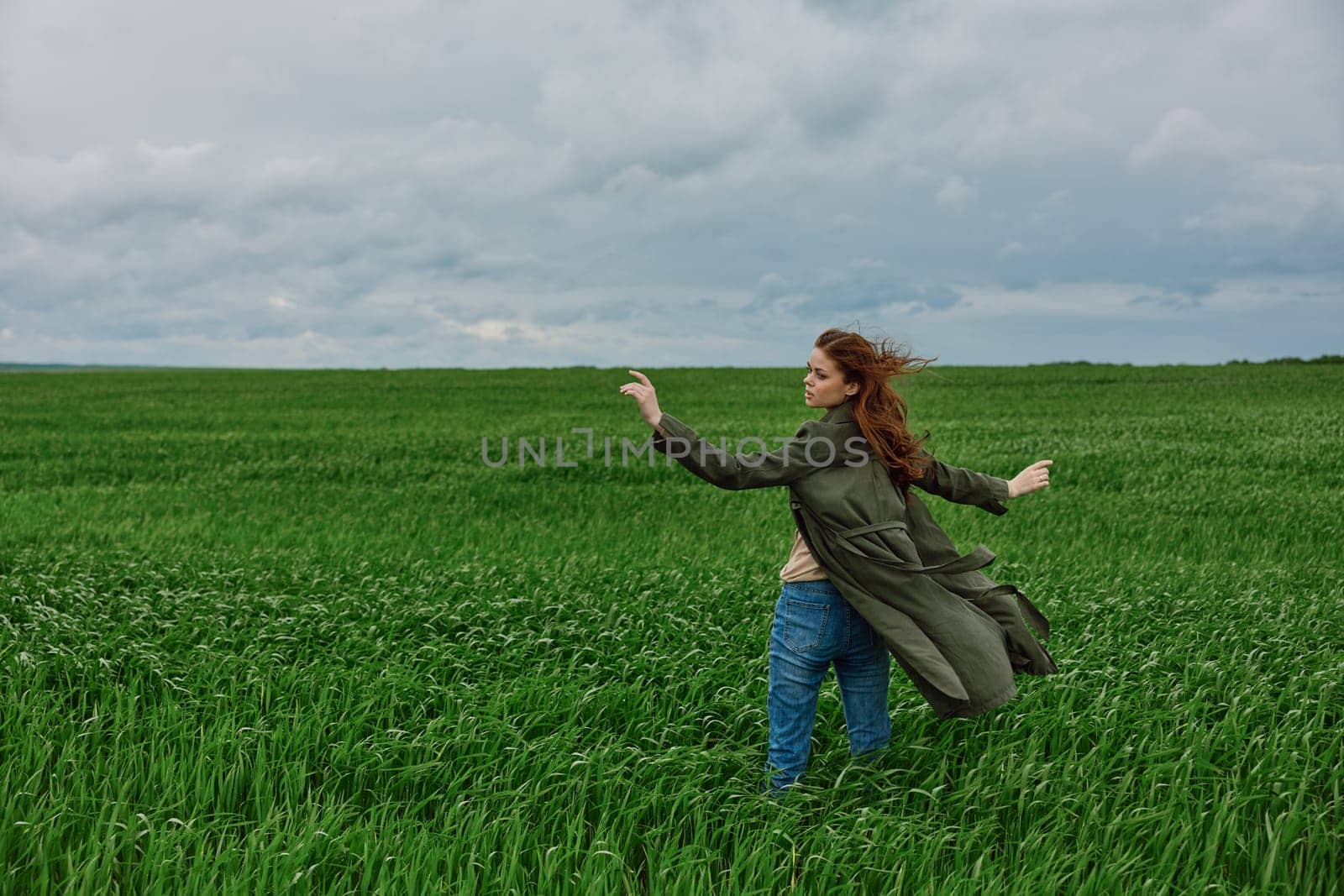 a woman in a long coat stands in tall green grass in a field, in cloudy weather, enjoying nature and the view by Vichizh