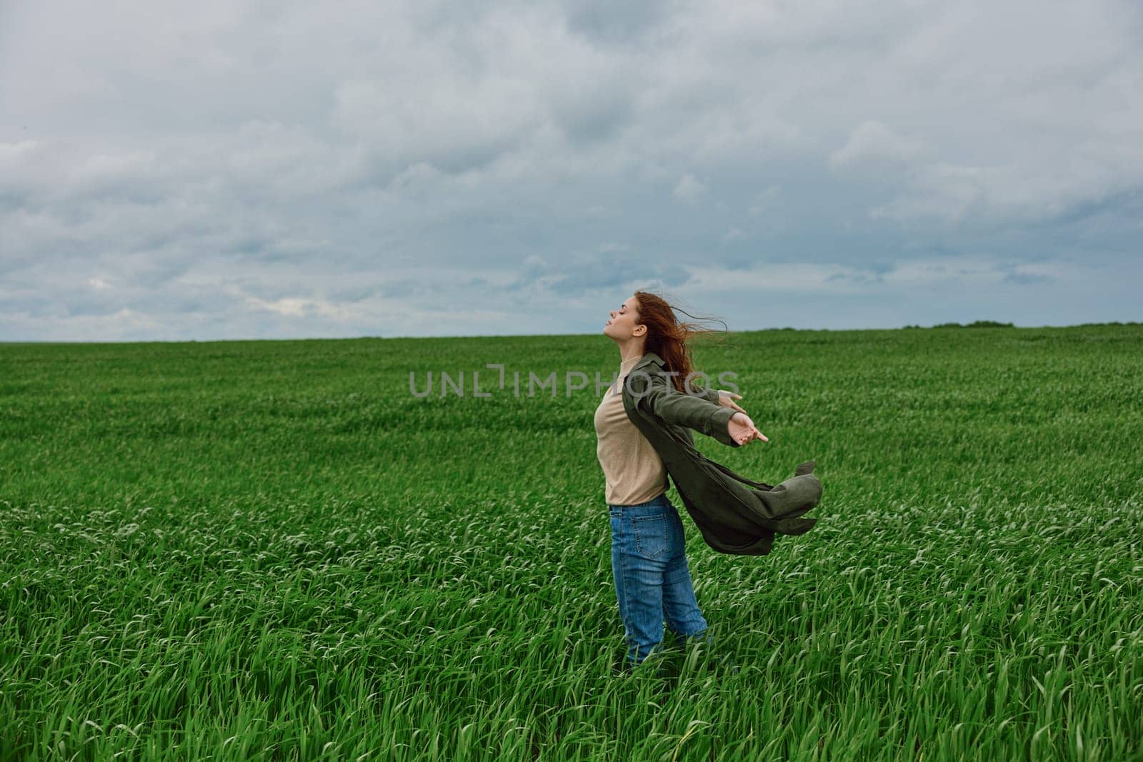 a woman in a long coat stands in tall green grass in a field, in cloudy weather, enjoying nature and the view by Vichizh