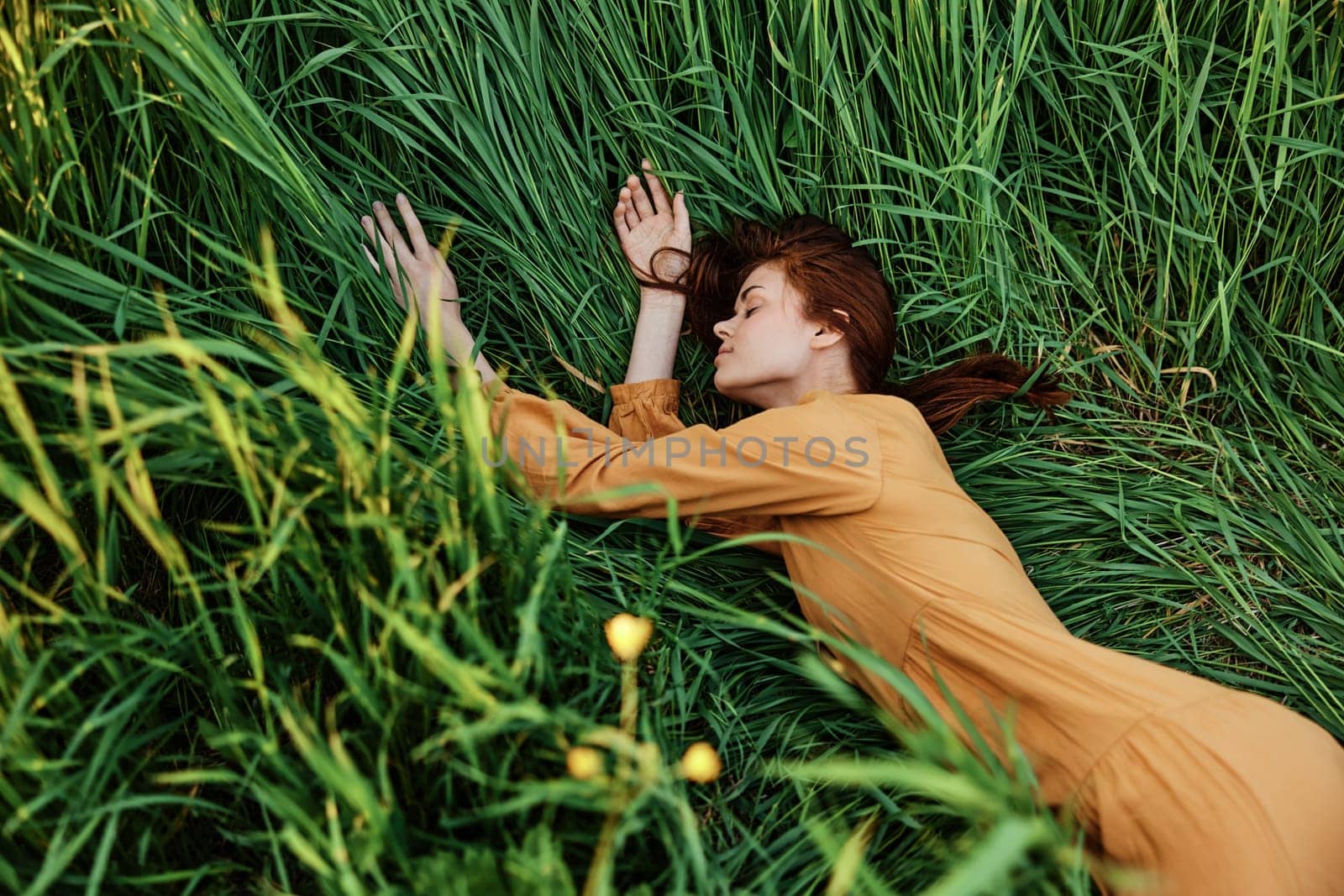 a close horizontal photo of a pleasant woman in a long orange dress resting lying in the tall grass with her eyes closed in sunny weather with her arms outstretched. High quality photo