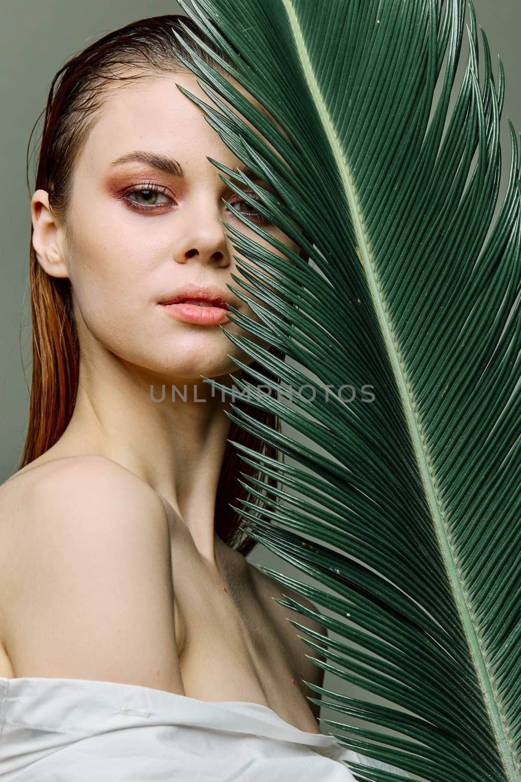 a sophisticated, elegant woman with evening makeup, stands with a green palm leaf, covering part of her face with it. Close vertical portrait, photo without retouching by Vichizh