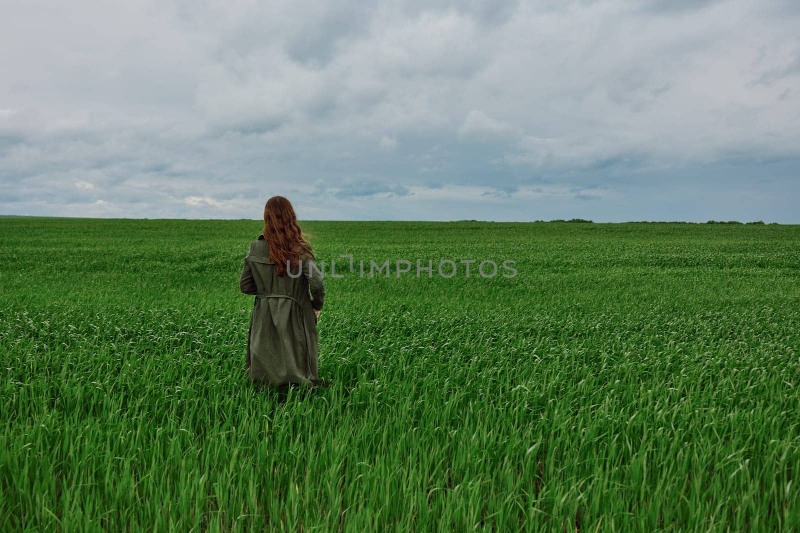 a woman in a long raincoat stands with her back to the camera, far away in a field in rainy, cloudy weather by Vichizh