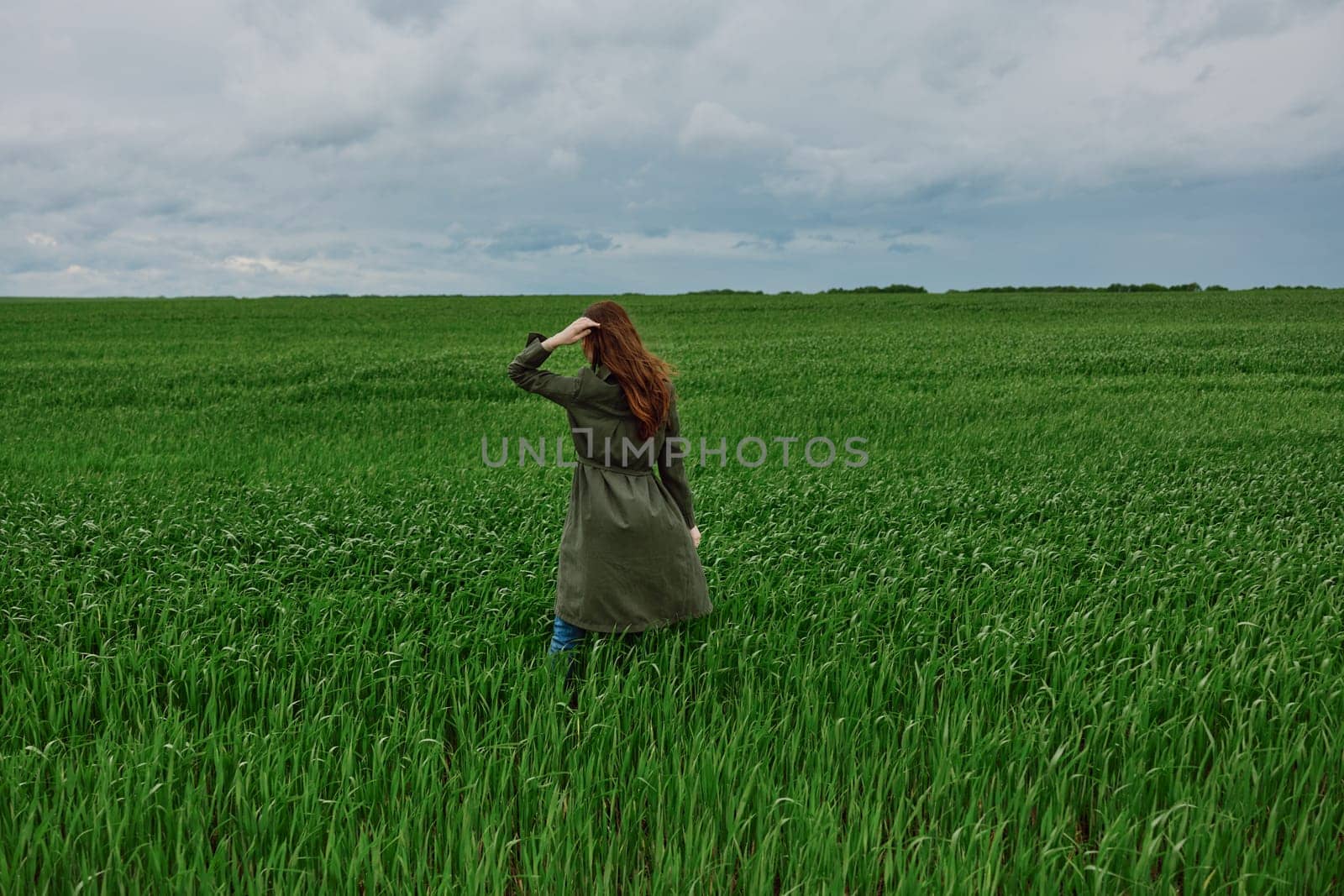 a woman in a long raincoat stands with her back to the camera, far away in a field in rainy, cloudy weather. High quality photo