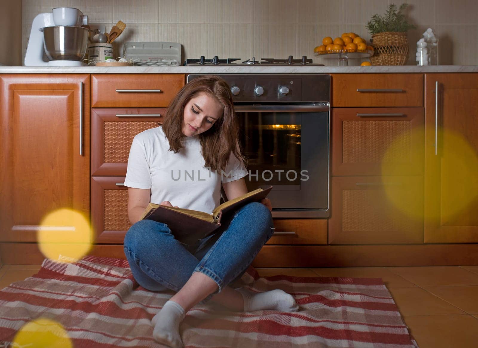 Horizontal shot of a happy woman sitting on the floor in the kitchen with an open notepad waiting for a delicious pie baking in the oven by aprilphoto