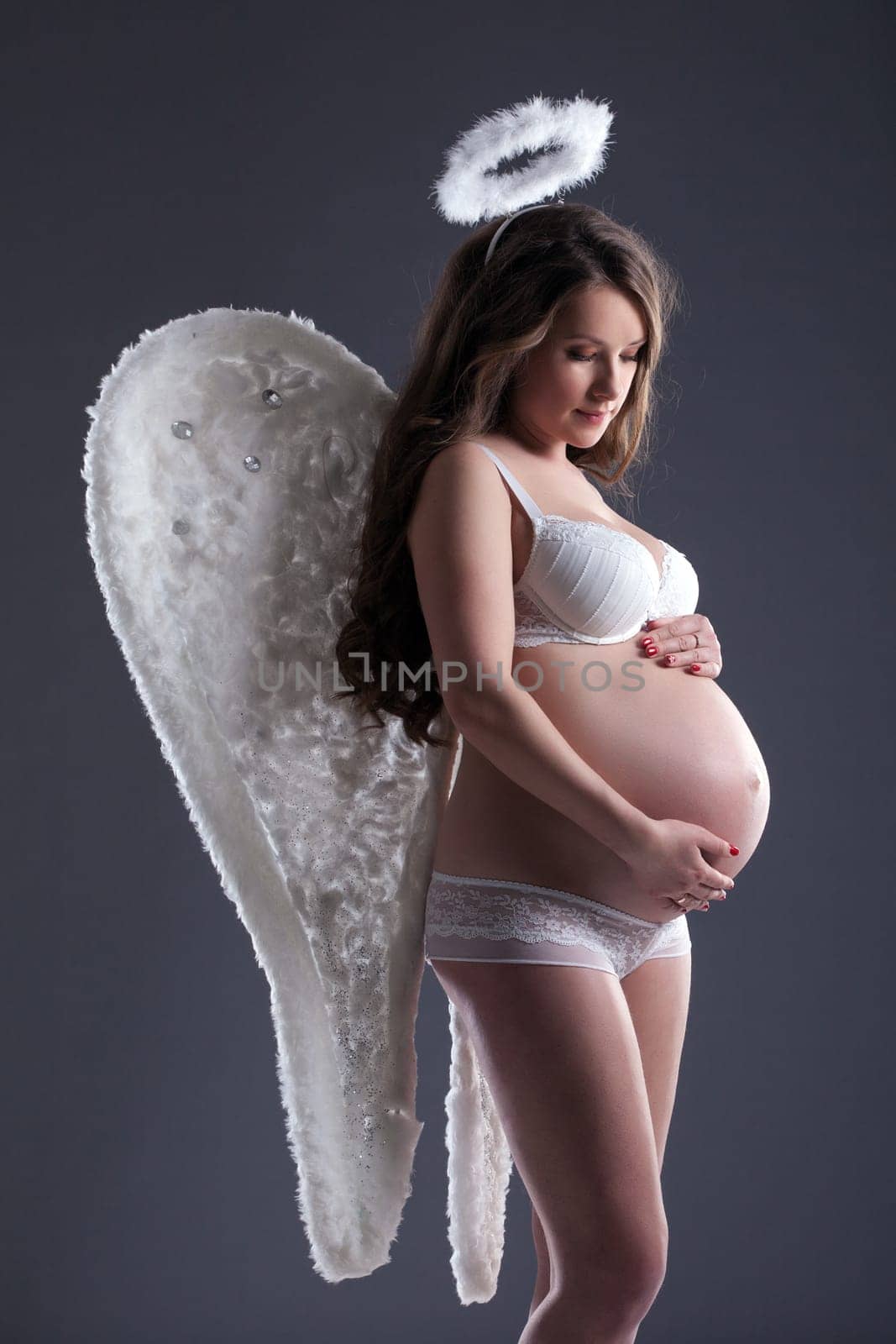 Pretty pregnant woman in angel costume posing on gray background