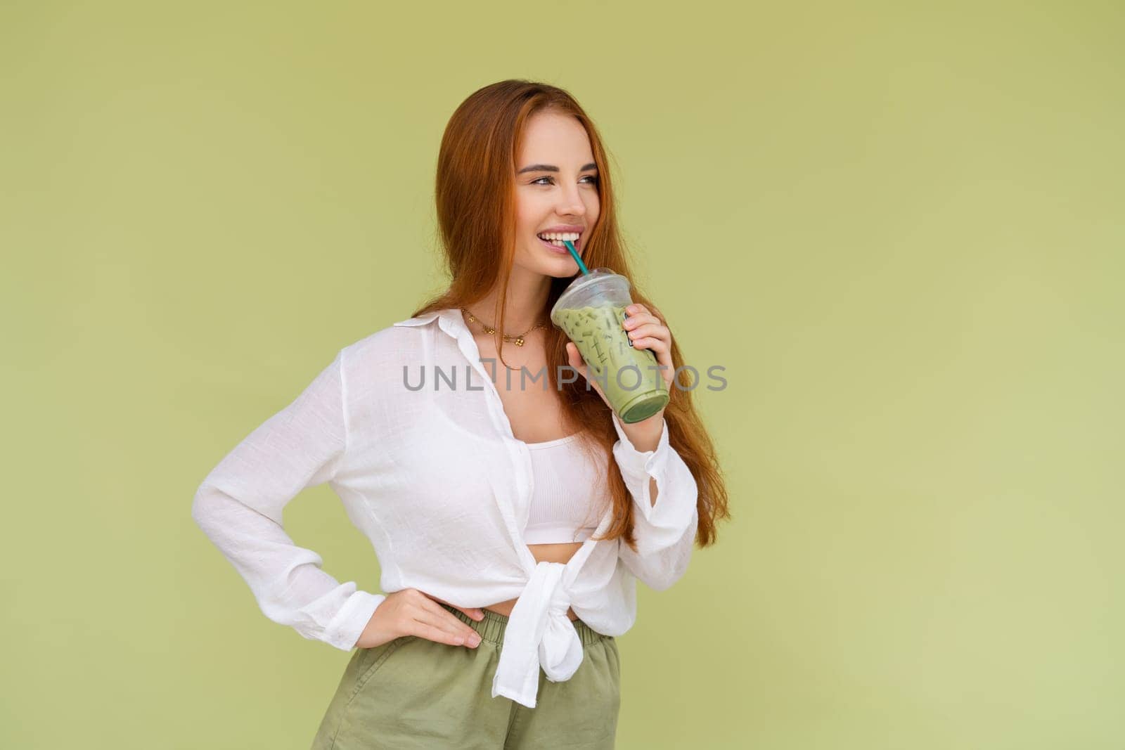 Beautiful red hair woman in casual shirt on green background positive vibes enjoying fresh ice green matcha tea with milk at hot summer day by kroshka_nastya