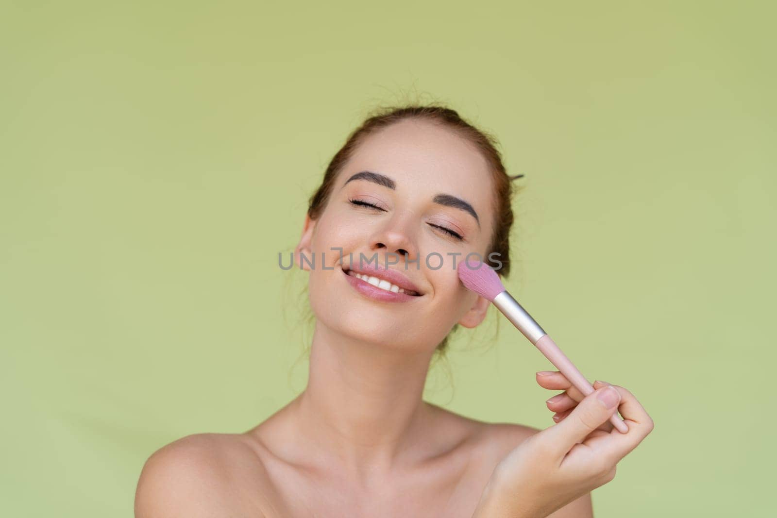 Beauty portrait of young topless red hair woman with bare shoulders on green background with perfect skin and natural makeup holds blush brush