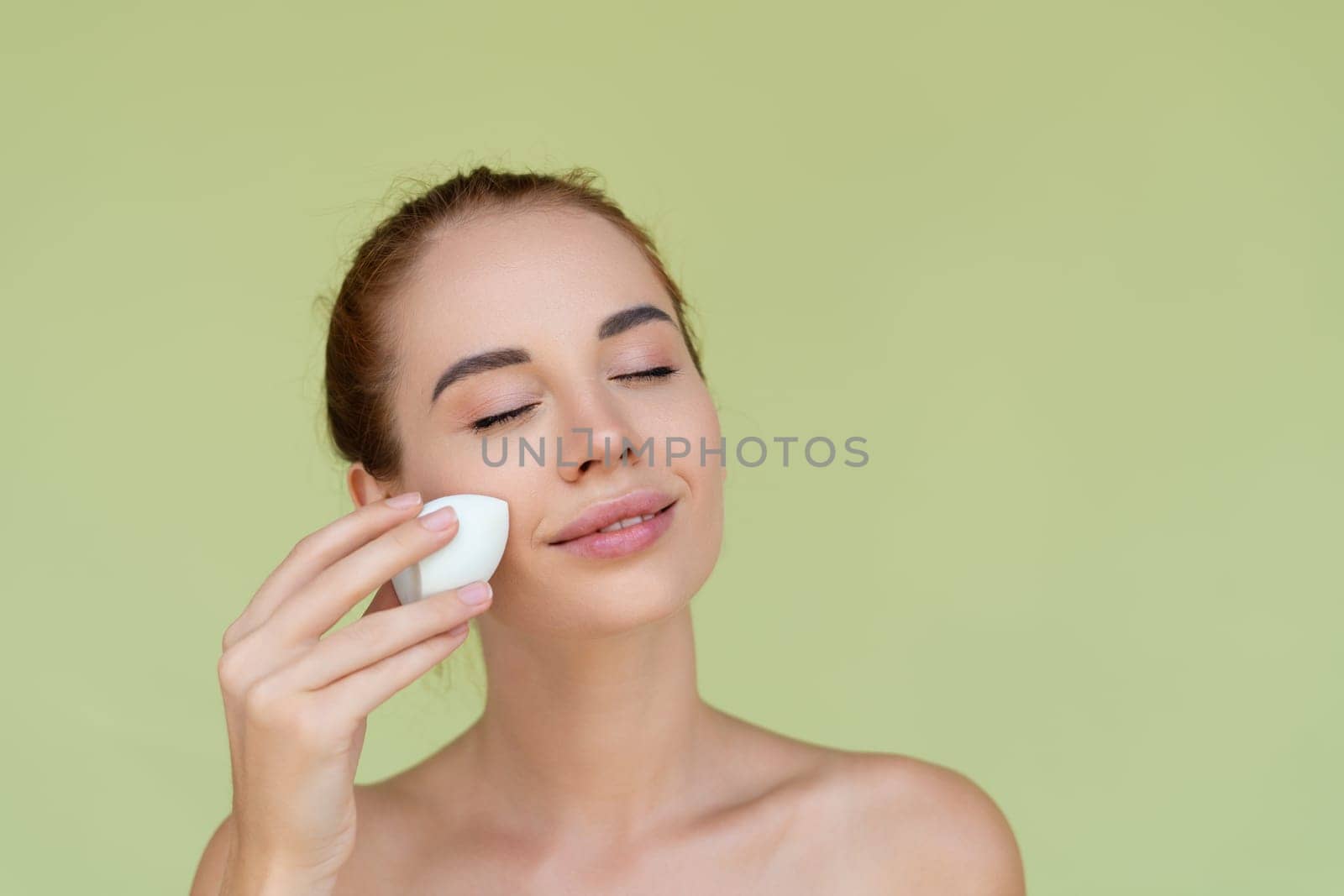 Beauty portrait of young topless red hair woman with bare shoulders on green background with makeup foundation sponge