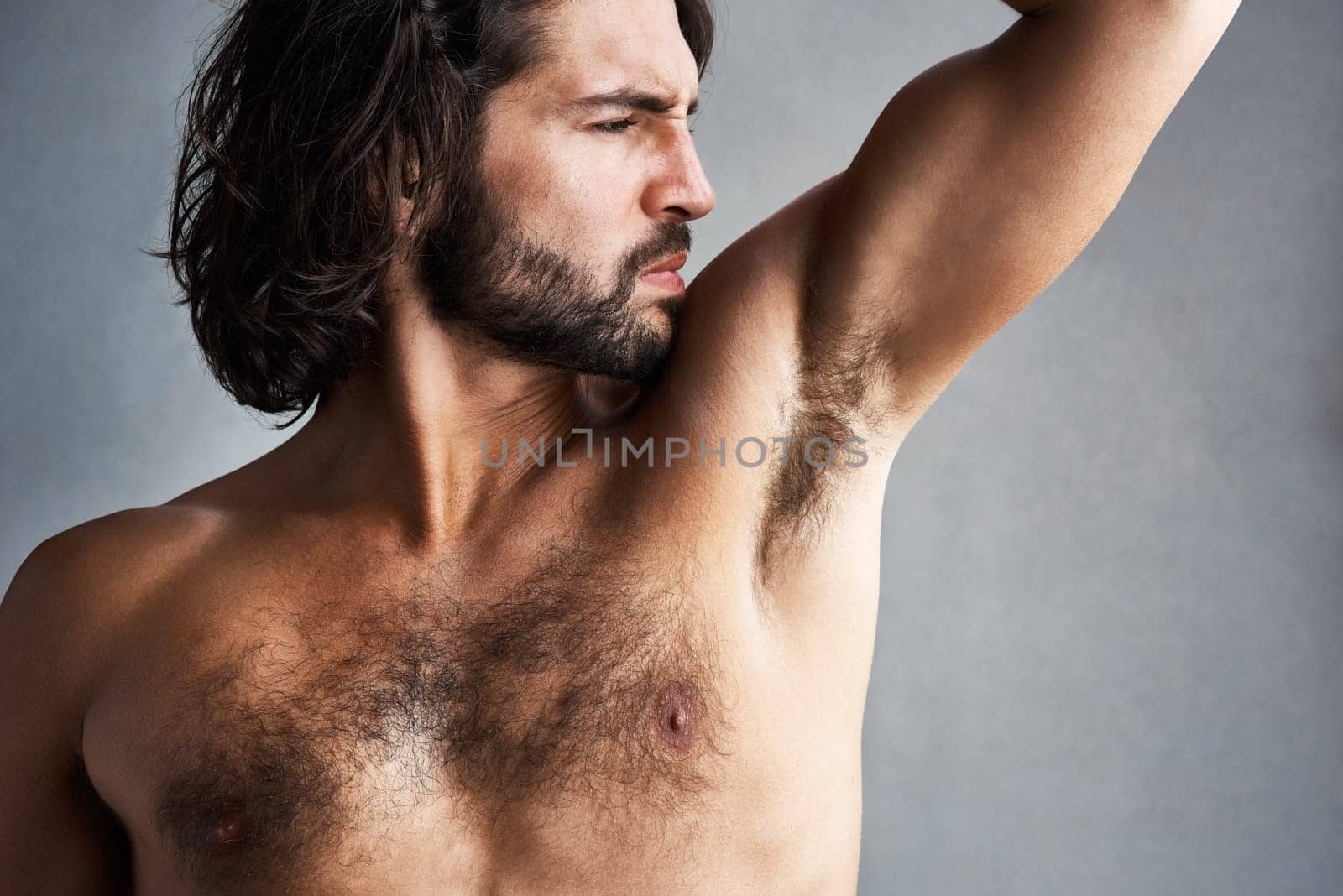 Body odour is never cool. Studio shot of a young man smelling his armpits against a grey background. by YuriArcurs