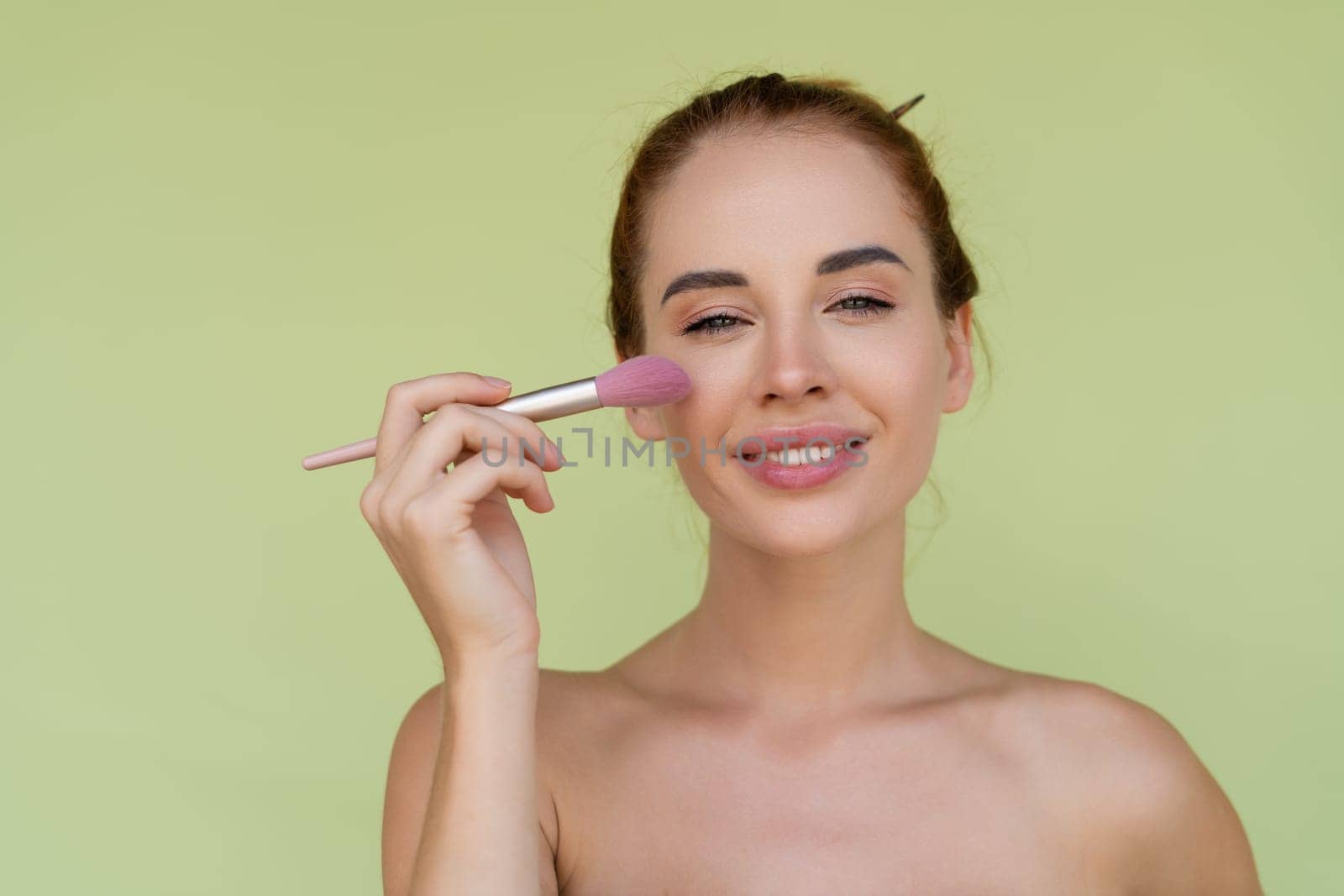 Beauty portrait of young topless red hair woman with bare shoulders on green background with perfect skin and natural makeup holds blush brush