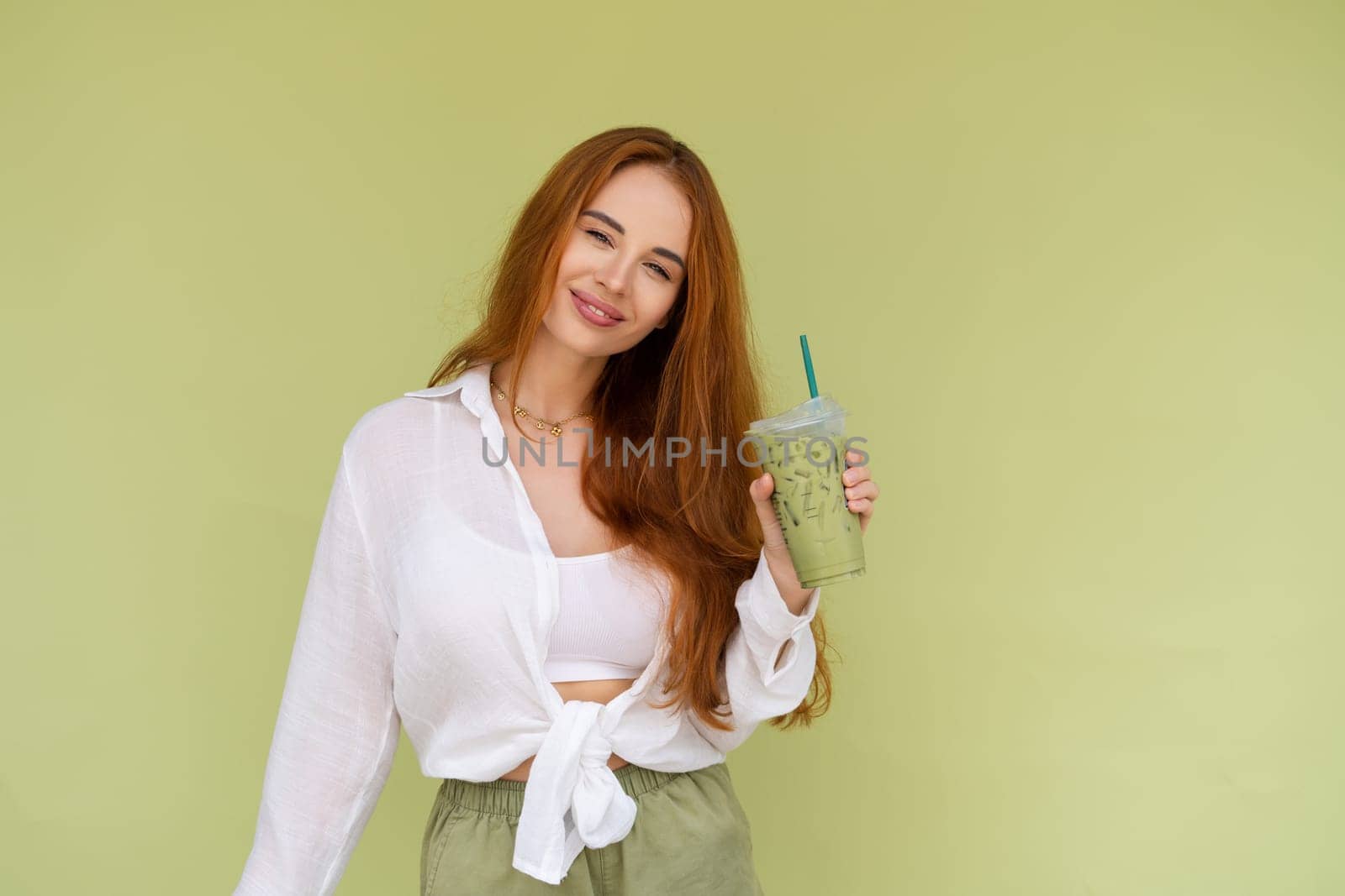 Beautiful red hair woman in casual shirt on green background positive vibes enjoying fresh ice green matcha tea with milk at hot summer day by kroshka_nastya