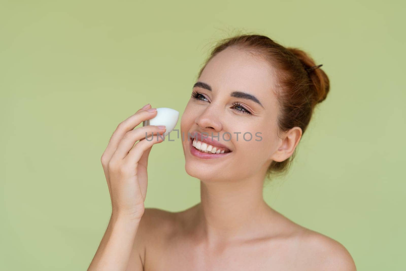 Beauty portrait of young topless red hair woman with bare shoulders on green background with makeup foundation sponge