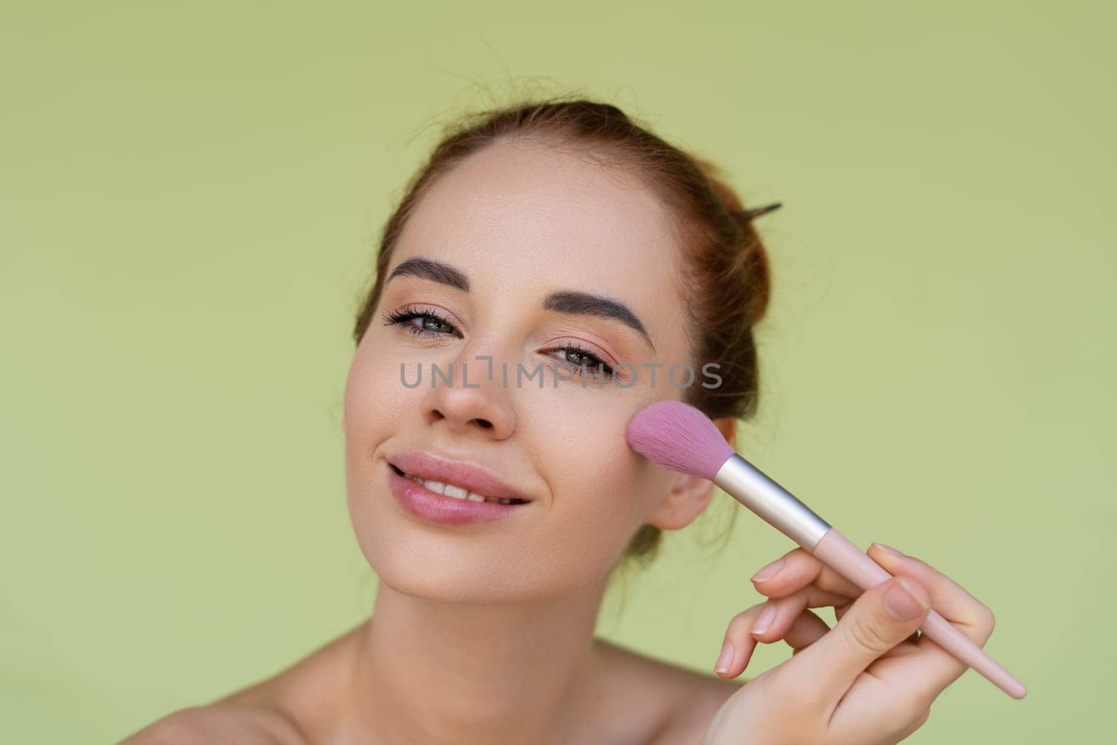 Beauty portrait of young topless red hair woman with bare shoulders on green background with perfect skin and natural makeup holds blush brush