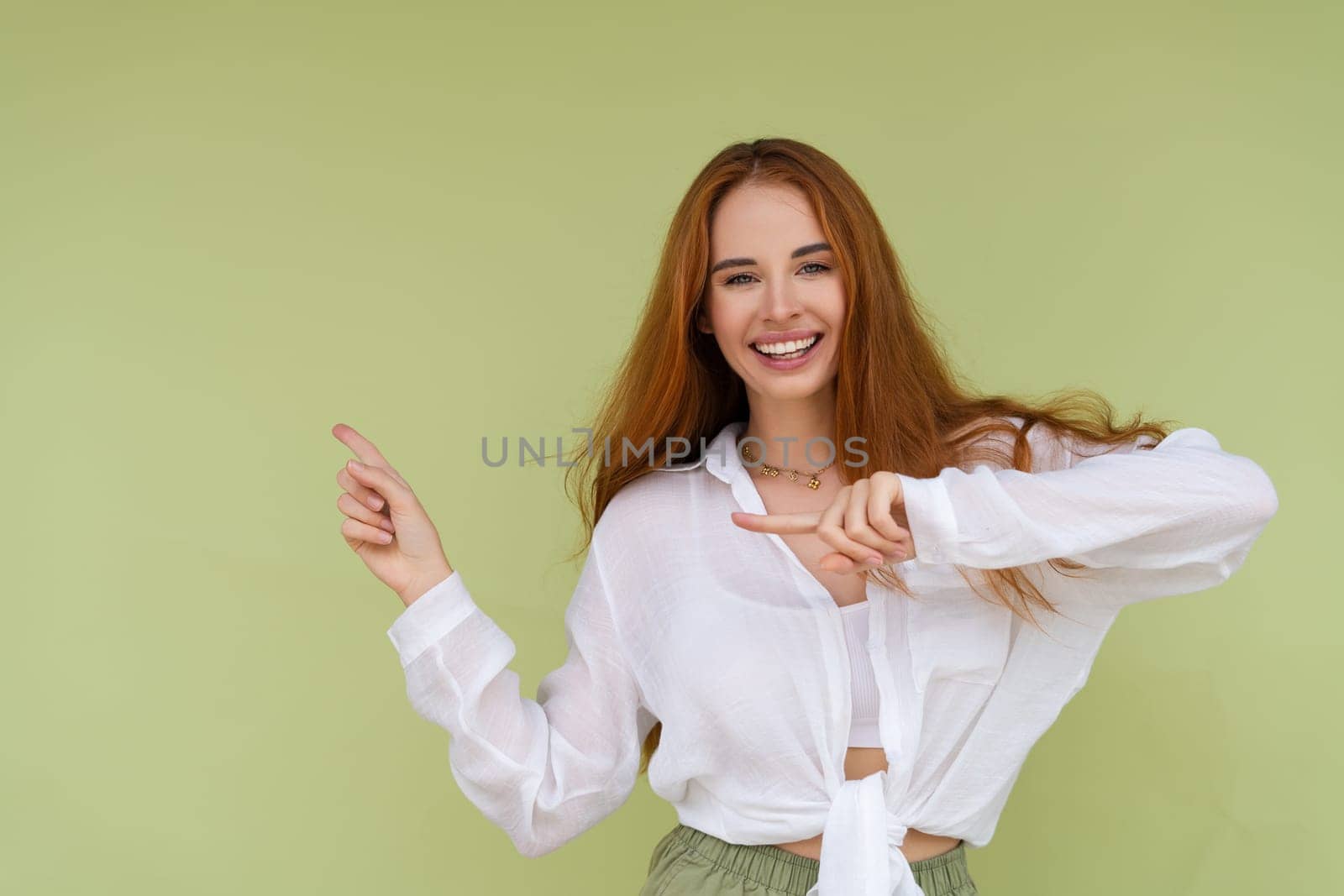 Beautiful red hair woman in casual shirt on green background happy look to camera smile excited point finger left
