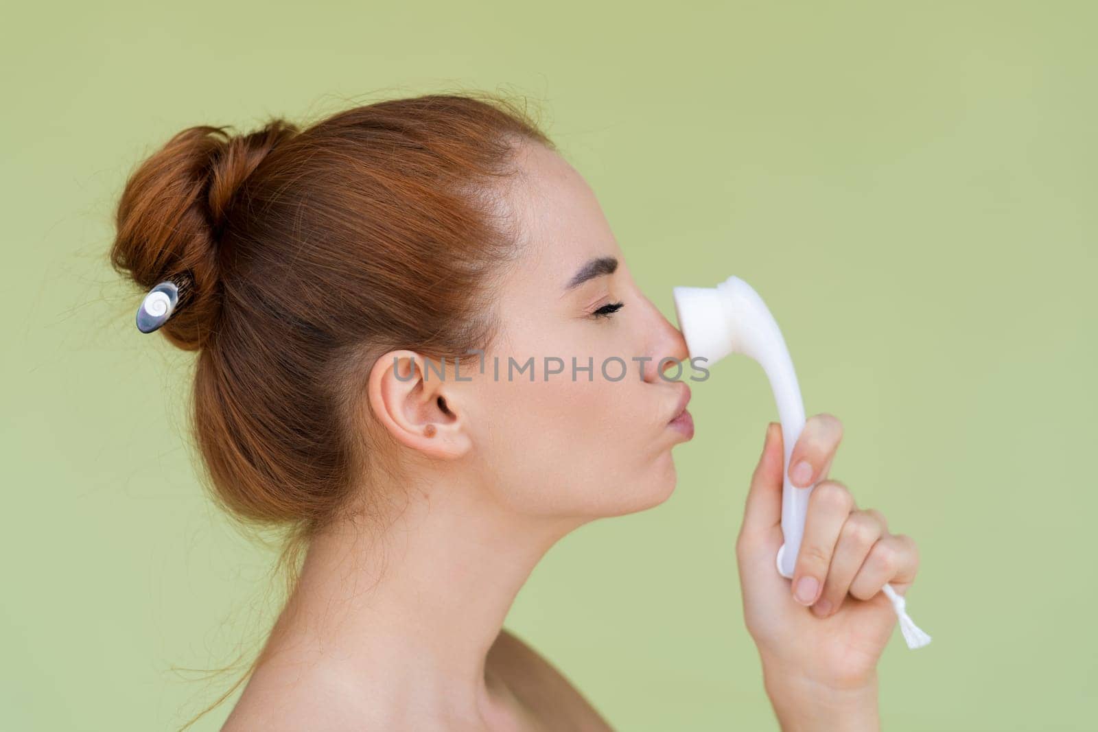 Beauty portrait of young topless red hair woman with bare shoulders on green background with perfect skin and natural makeup holds face cleansing brush