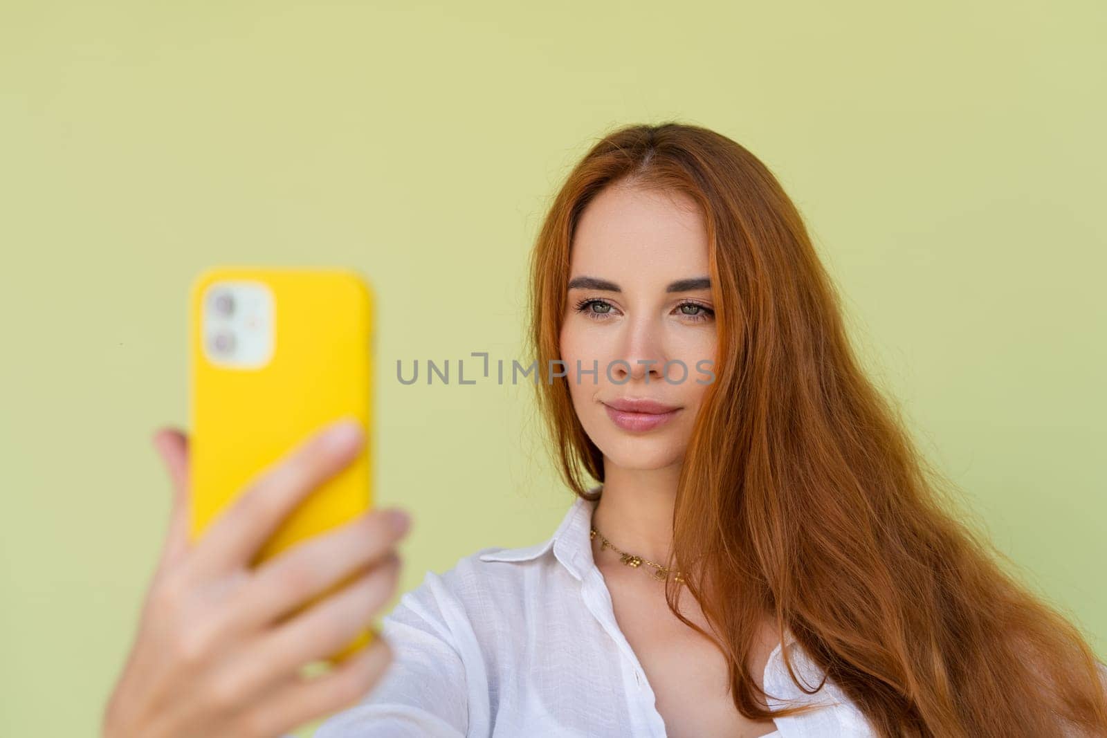 Beautiful red hair woman in casual shirt on green background happy taking photo selfie on smart phone by kroshka_nastya