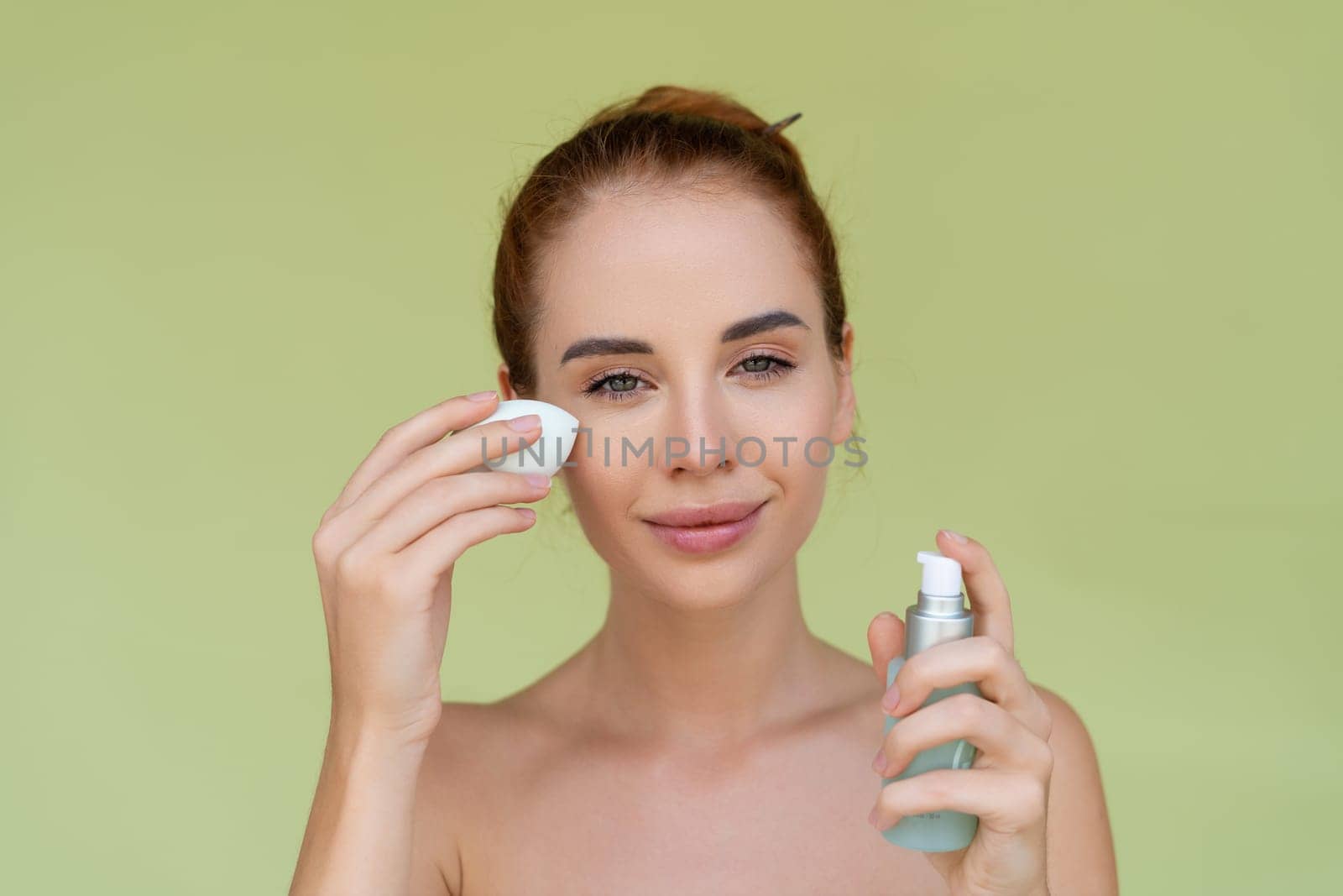 Beauty portrait of young topless red hair woman with bare shoulders on green background holds serum for youth and skin hydration and sponge