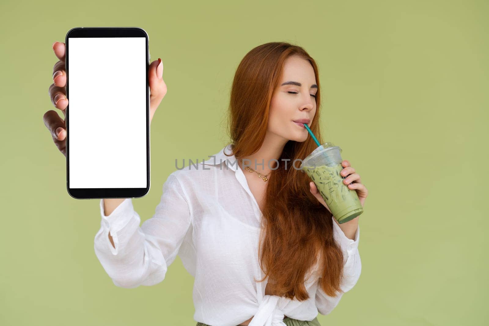 Beautiful red hair woman in casual shirt on green background holds a phone with a blank white screen and matcha ice green tea latte by kroshka_nastya