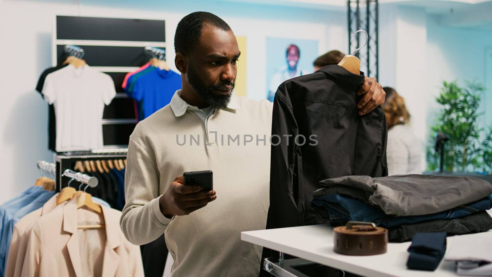 Young client analyzing online website app to find clothes, buying new fashion collection in department store. Customer with mobile phone checking merchandise of formal wear, small business.