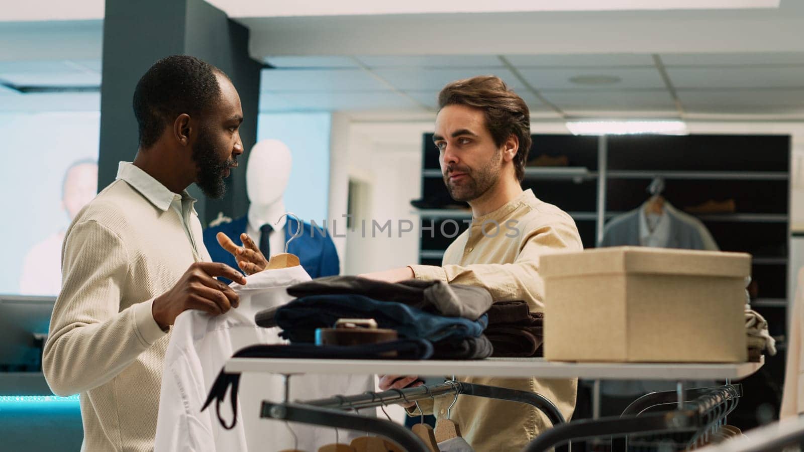 Store manager helping client to choose clothes from hangers, young man checking formal wear brands for commercial activity. African american customer looking at shop merchandise.