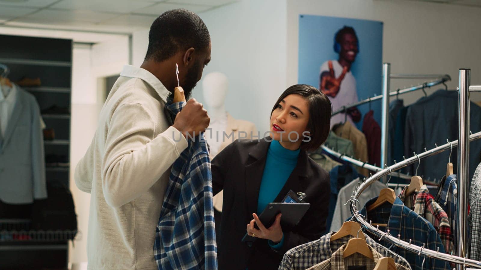 Asian employee giving shirts to client to try on in clothing store, helping customer to buy modern clothes. Woman talking to young man about new fashion collection, small business. Handheld shot.