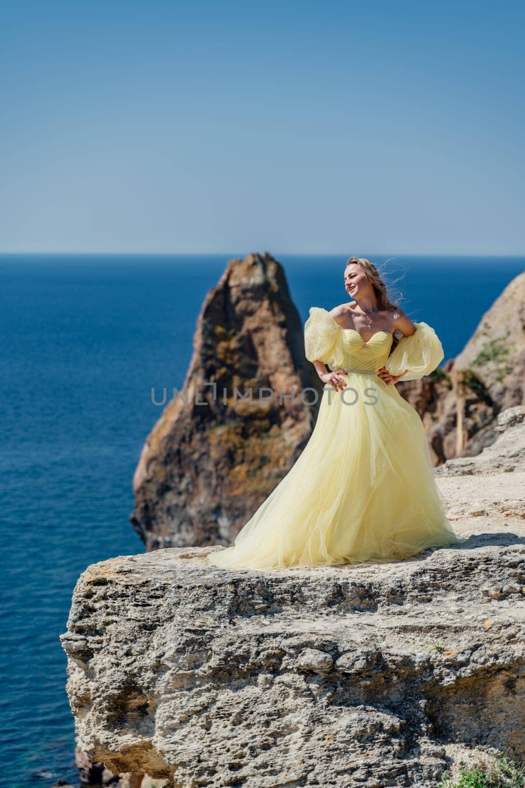 Woman in a yellow dress on the sea. Side view Young beautiful sensual woman in yellow long dress posing on a rock high above the sea at sunset. Girl in nature against the blue sky by Matiunina