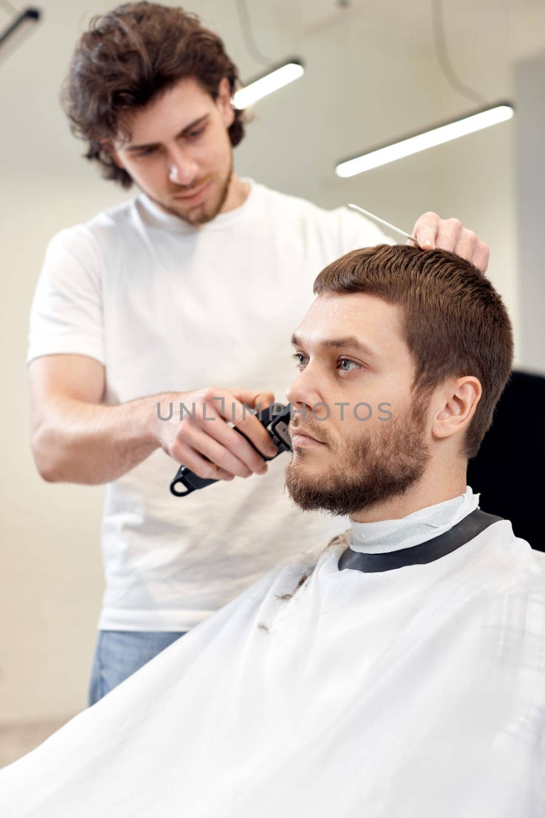 Barber trim hair with clipper on handsome bearded man in barber shop.