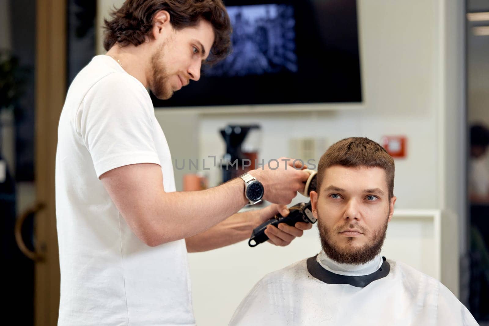 Barber trim hair with clipper on handsome bearded man in barber shop.