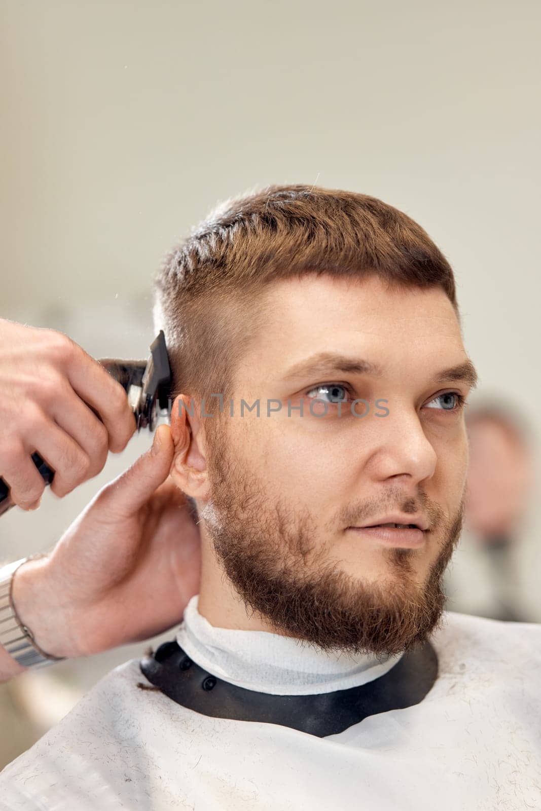 Barber trim hair with clipper on handsome bearded man in barber shop.