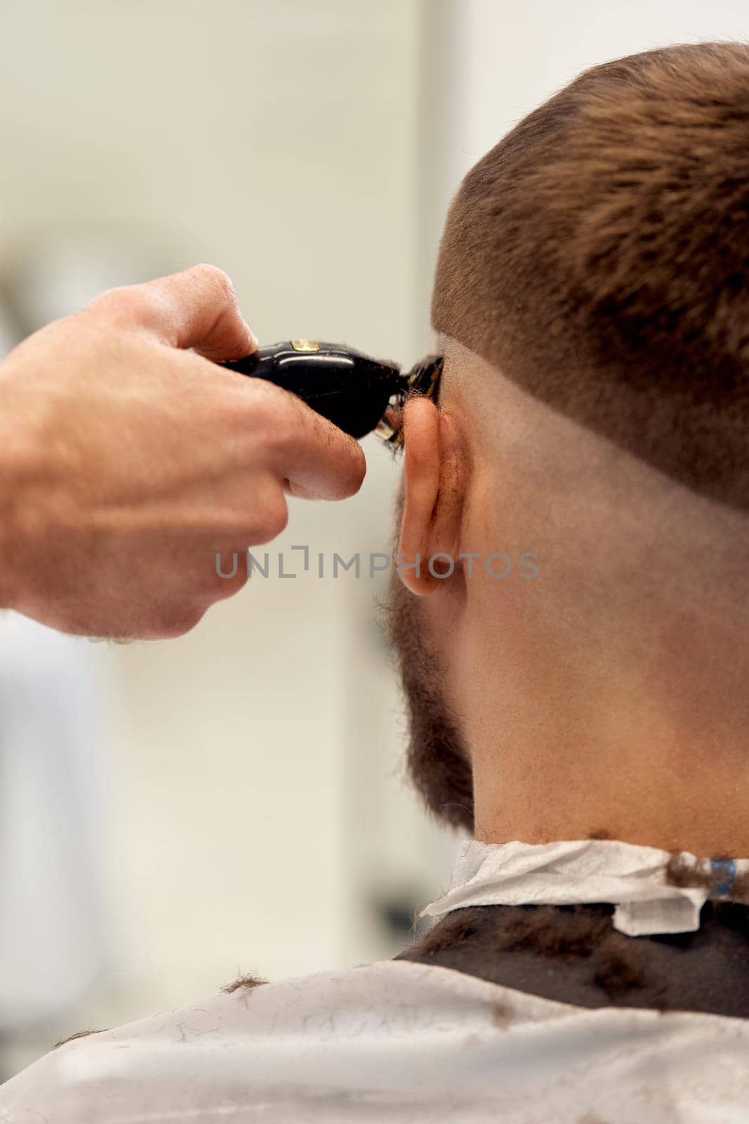 Barber trim hair with clipper on handsome bearded man in barber shop.