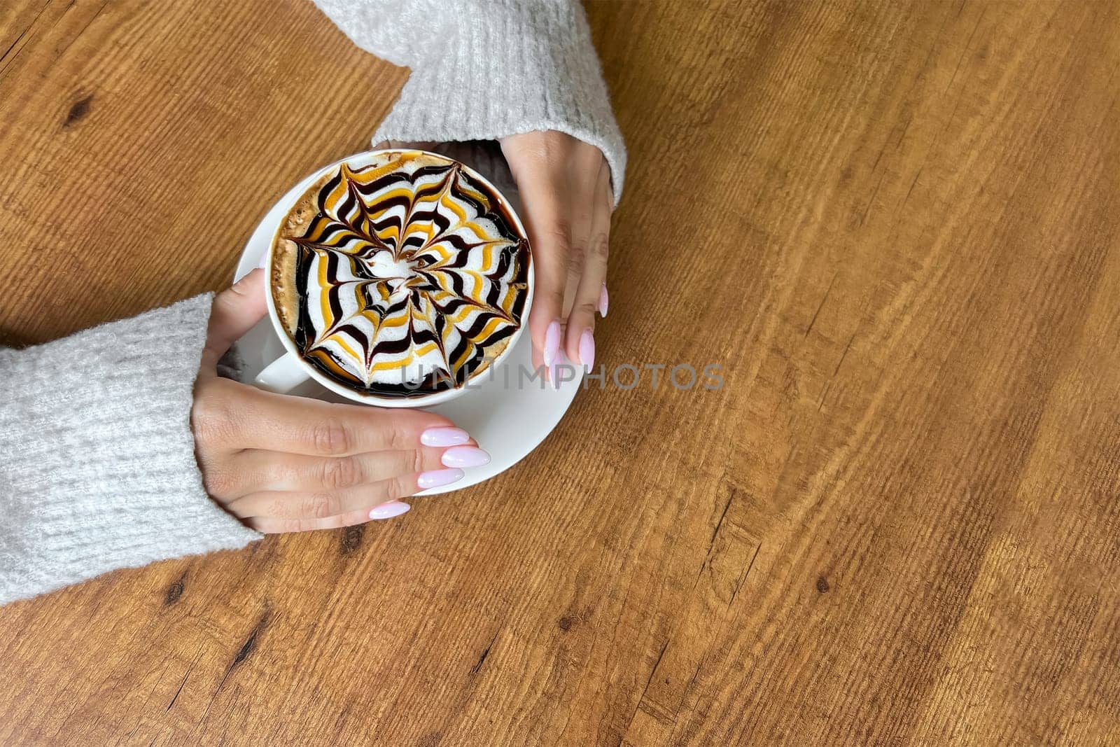 Hands of girl hold white cup of coffee with spider web pattern on top of foam, against background of wooden table, copy space, top view