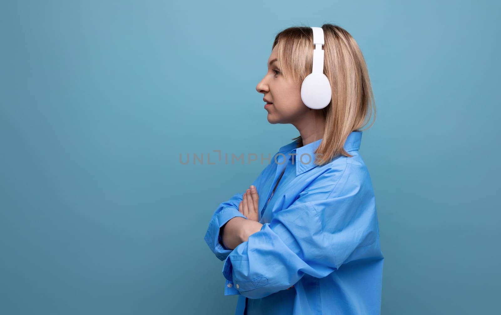 horizontal profile photo of an adorable young woman in a casual shirt focused on sound in large white headphones on a blue isolated background with copy space.