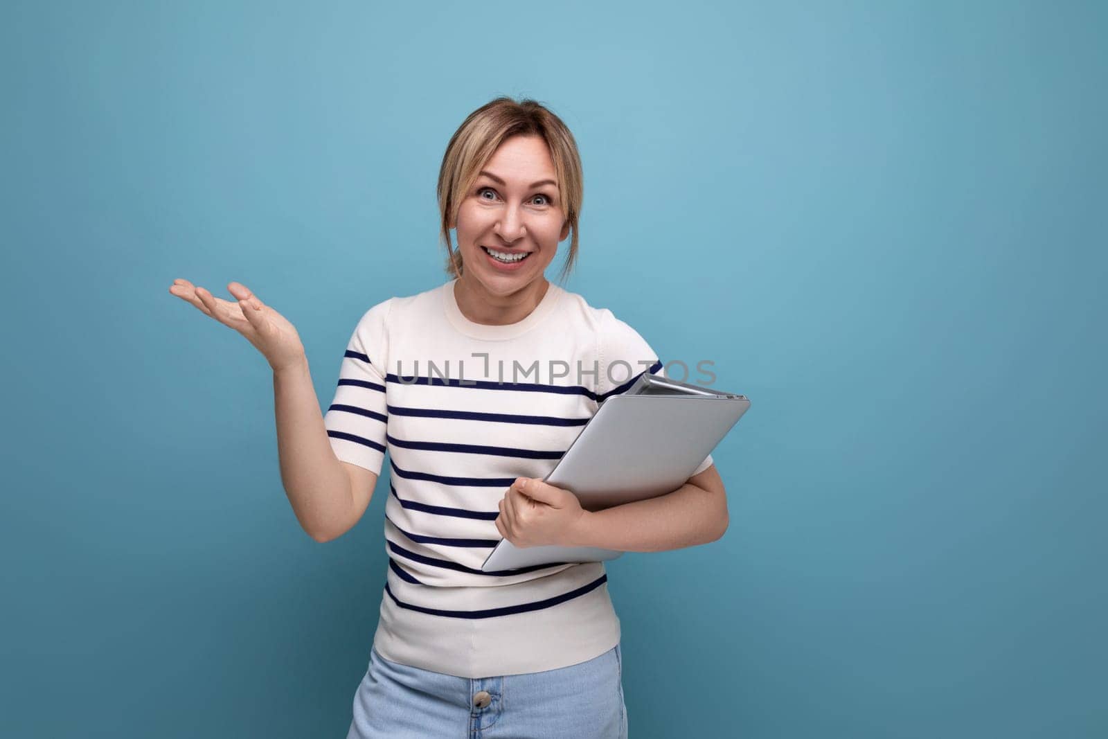 close-up of a successful blonde woman freelancer in a striped sweater holding a laptop in her hands on a blue background.