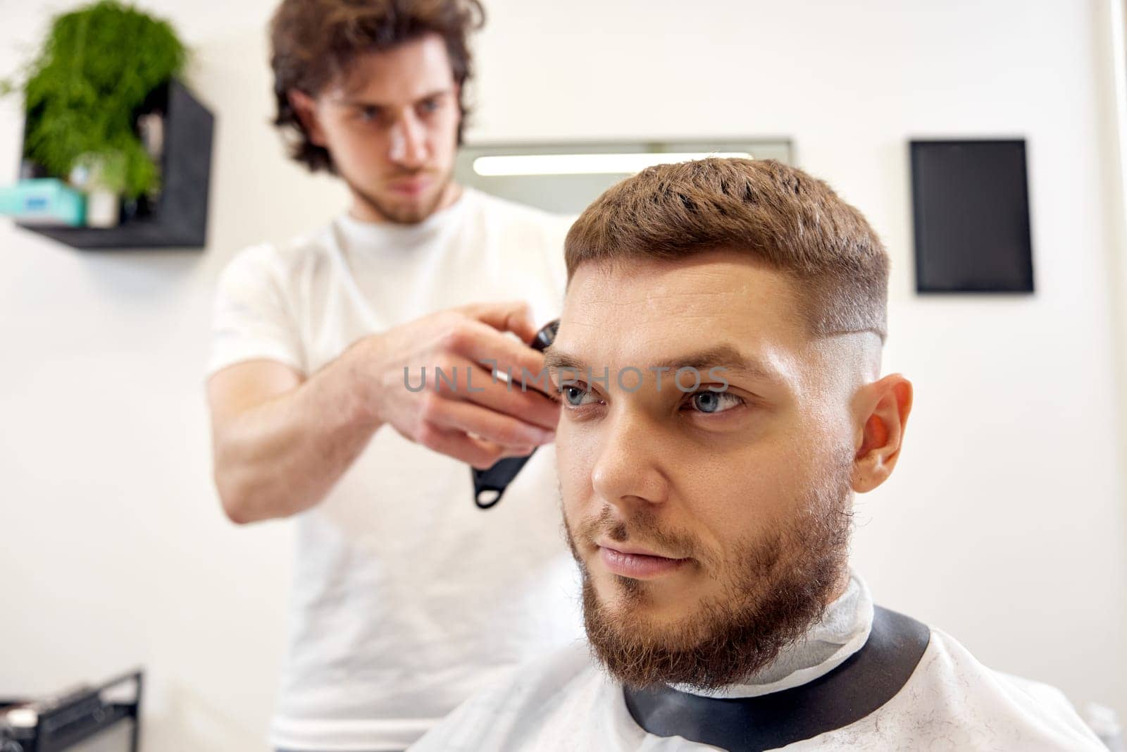 Barber trim hair with clipper on handsome bearded man in barber shop.