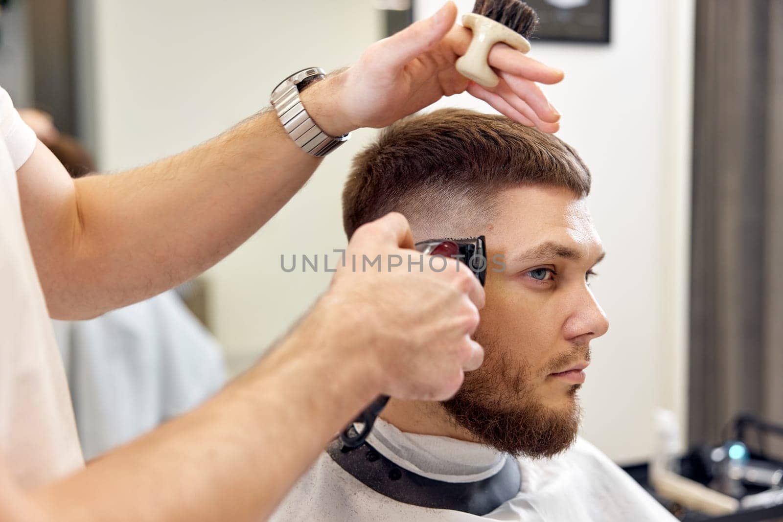 Barber trim hair with clipper on handsome bearded man in barber shop.